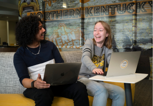 A male and a female student laughing and sitting in front of their laptops